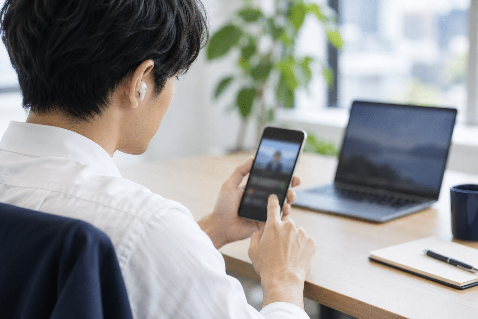 明るいオフィスでイヤホンを着けてスマートフォンの動画音声を選ぶ人